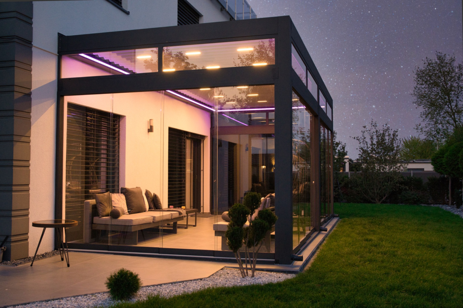 Modern glass-walled patio at night with illuminated purple LED lights, outdoor seating with pergola system, and a view of the starry sky and garden.