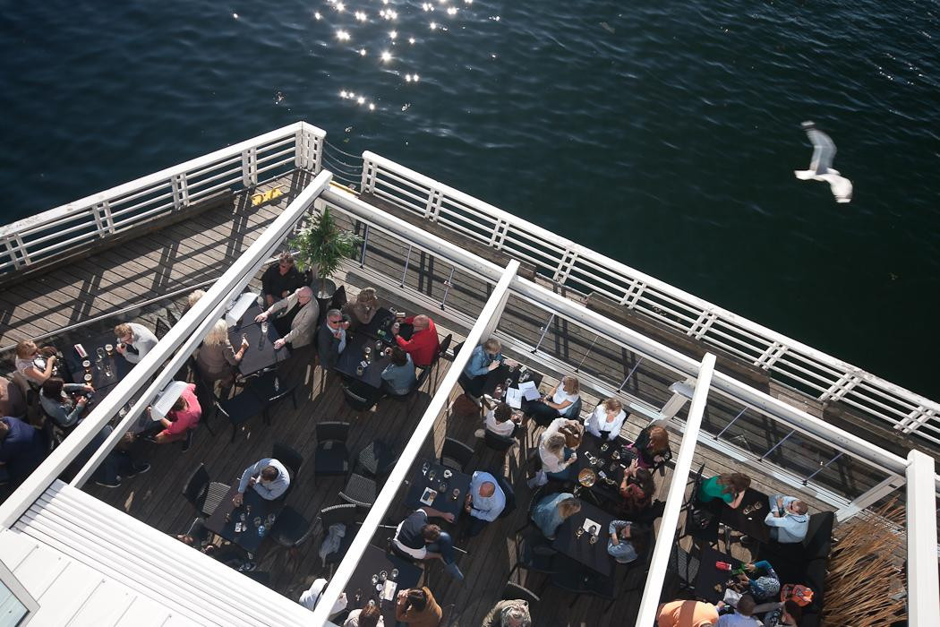 A lively group enjoys dining at tables on a deck, with a scenic sea view, enhancing customer engagement in a pergola setting.