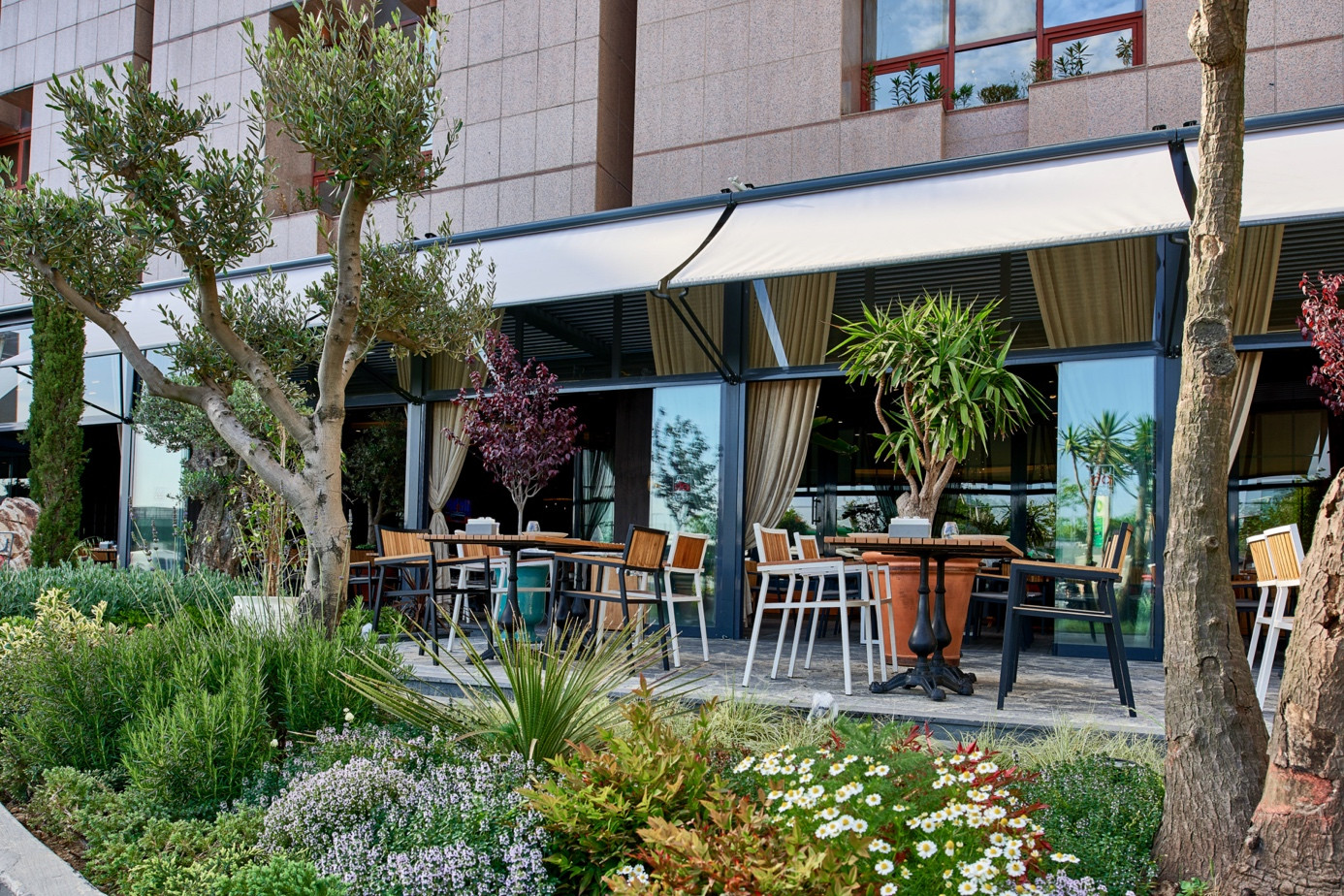 Façade-compatible aluminum awning system designed for a modern restaurant terrace with landscaped outdoor seating