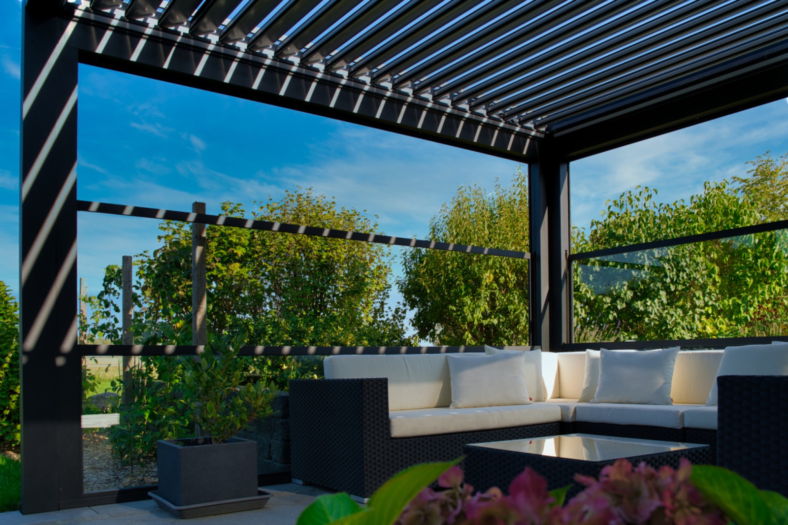 Lamelli pergola tavanı ve cam yan panellerle çevrili, bahçeye bakan oturma alanı.